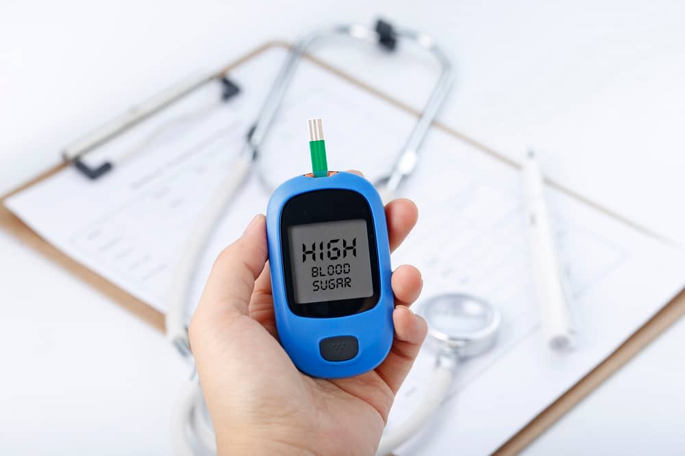 Hand holding a blood glucose meter measuring blood sugar, the background is a stethoscope and chart file